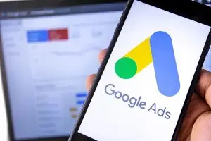 Hand holding a smartphone showing the Google Ads logo, with a blurred laptop displaying an ads dashboard behind it.