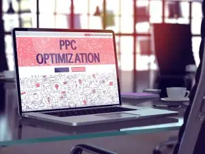 Laptop on a glass office desk displaying a pink doodle-style landing page headed "PPC Optimization", with a coffee cup nearby