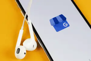 Smartphone on a yellow background showing the blue Google My Business storefront logo, with white wired earbuds beside it.
