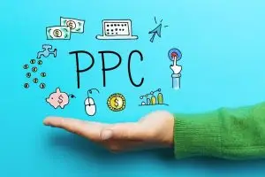 Open palm in a green sweater against bright blue, holding doodled "PPC" lettering with banknotes, a laptop, piggy bank, coins and a click cursor