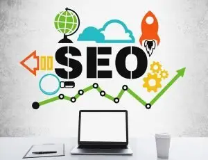 Laptop, clipboard and coffee cup on a white desk before a concrete wall painted with "SEO", a rocket, globe, gears and a rising green line chart