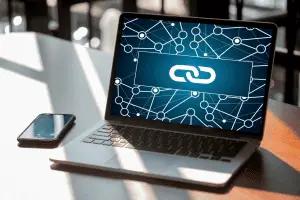Laptop open on a sunlit white desk next to a phone, its screen showing a white chain-link icon over a dark blue network of connected nodes