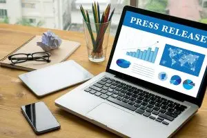 Laptop on a wooden desk showing a blue "Press Release" dashboard of charts, beside a notebook, glasses, colored pencils, tablet and phone
