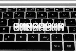 Black-and-white close-up of white letter dice spelling "Keywords" twice, resting on a laptop keyboard