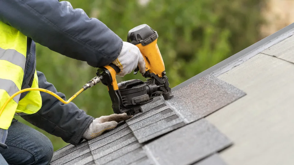 Roofing contractor in a safety vest nailing asphalt shingles with a pneumatic nail gun